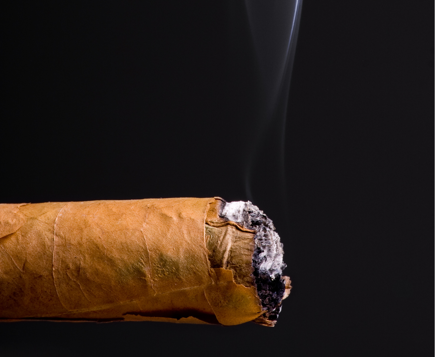 Nahaufnahme einer rauchenden Zigarre vor schwarzem Hintergrund. Der Körper der Zigarre ist hellbraun und wird nach rechts hin dünner. Am linken Ende befinden sich Asche und Rauch. Der Rauch steigt in dünnen Linien nach oben.